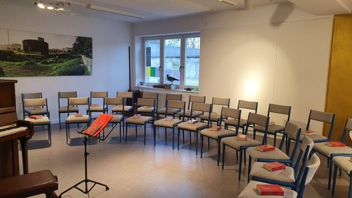 Ein Raum mit Stühlen und einem Klavier, ein Bild an der Wand, ein Fenster mit Blick auf Bäume.