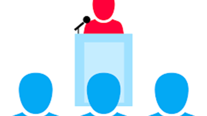 Person taler ved talerstol, mens tre andre sidder ved bord