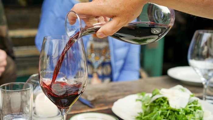 En person serverer rødvin i glas på spisebord med mad og andet glasvin.