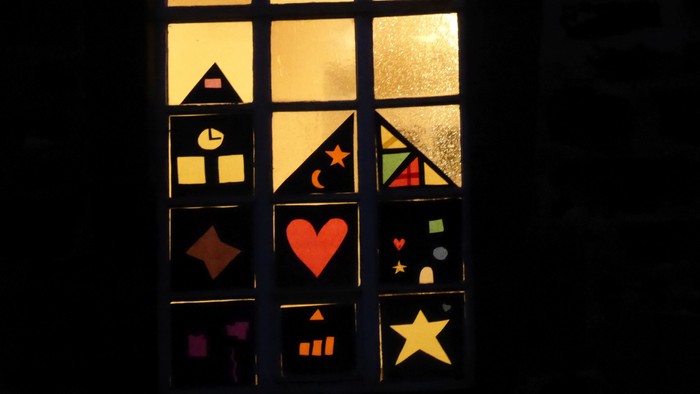 Ein Fenster mit bunten geometrischen und herzförmigen Ausschnitten, das nachts von hinten beleuchtet wird.