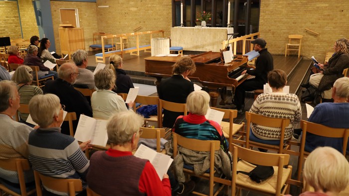 En gruppe mennesker deltager i en sangstund i et rum med en piano.