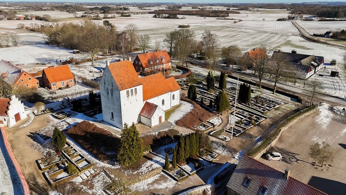 Luftfoto af en idyllisk landsbykirkegård omgivet af snedækkede marker og historiske bygninger med røde tage.