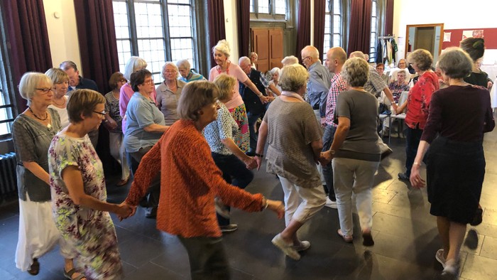 Eine Gruppe älterer Menschen tanzt in einem großen Raum mit hohen Fenstern.