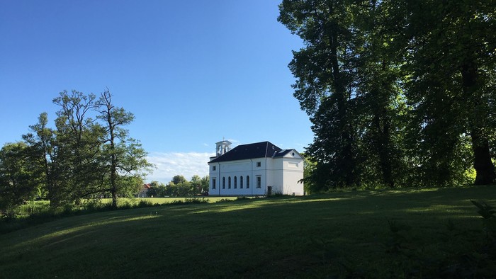 Et hvidt hus med sort tagtårn i en grøn og træfattig have under et klarblåt himmel.