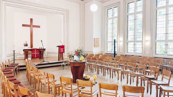 Auf dem Bild ist ein heller Raum zu sehen. Vorne stehen viele Holzstühle im Halbkreis. Hinten ist ein kleiner Altar mit einem großen Holzkreuz. Daneben steht ein Rednerpult mit einem roten Tuch. Der Raum hat große Fenster und viel Licht. In der Mitte steh