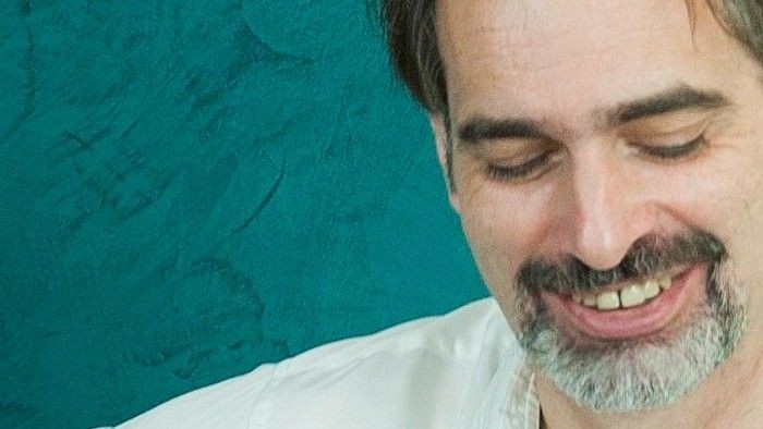 Ein lächelnder Mann mit Bart und Schnurrbart vor einem türkisfarbenen Hintergrund.