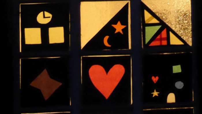 Ein Fenster mit bunten geometrischen und herzförmigen Ausschnitten, das nachts von hinten beleuchtet wird.