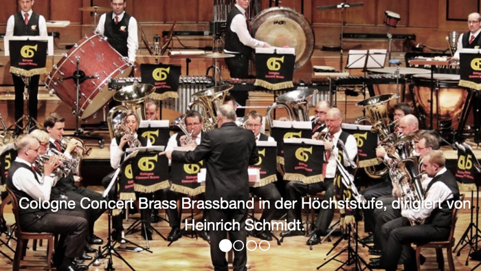 Ein Blechblasorchester, das in einem Konzertsaal unter der Leitung von Dirigent Heinrich Schmidt auftritt.
