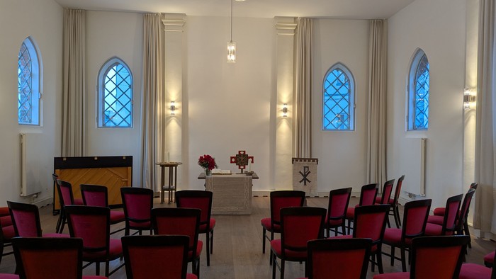 Ein heller Raum mit hohen Fenstern und roten Stühlen, ein Altar mit Kerzen und Blumen.