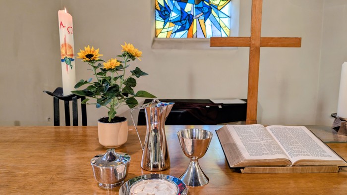 Auf dem Tisch stehen eine Vase mit Sonnenblumen, silberne Kelche und ein offenes Buch neben einem Holzkreuz