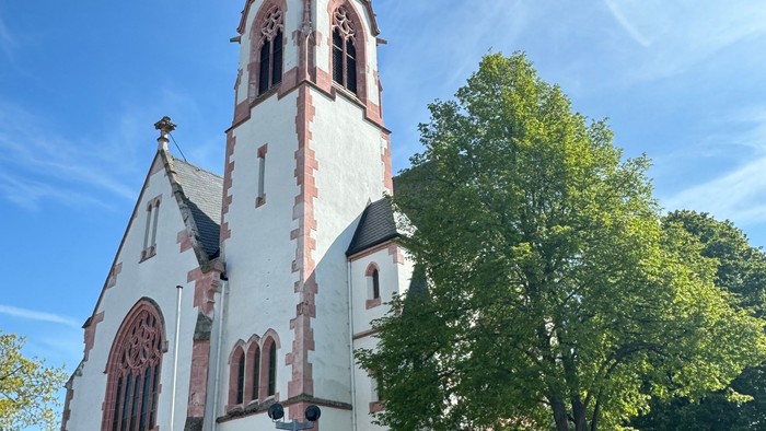 Weiße Kirche mit hohem Turm und spitzen Dach, umgeben von Bäumen und einem klaren blauen Himmel.