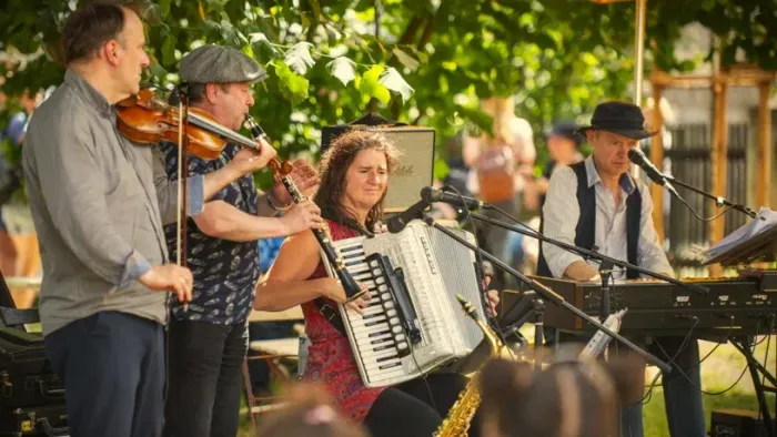 Eine lebhafte Freiluftband spielt traditionelle Musik mit Geigen, Akkordeon und Saxophon in einer grünen Parklandschaft.