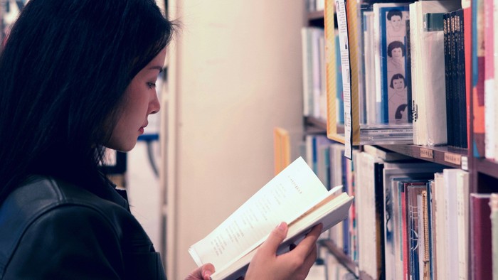 Eine Person liest ein Buch, während sie vor einer Wand steht, die mit verschiedenen Büchern gefüllt ist.