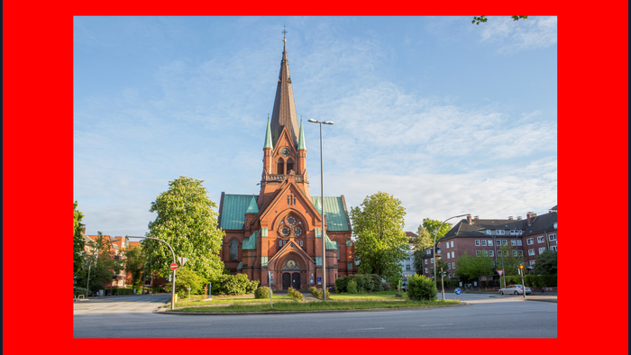 Ein rotes Backsteingebäude mit hohem Turm und grünem Dach, umgeben von Bäumen und einem klaren blauen Himmel.