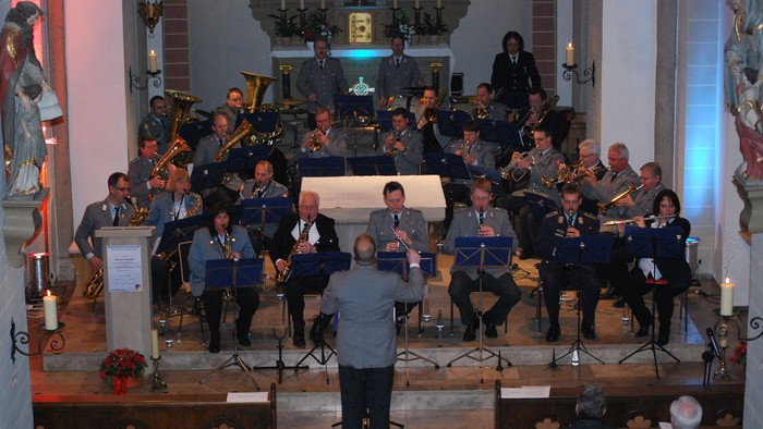 Eine Blaskapelle spielt in einer Kirche vor sitzendem Publikum, dirigiert von einem Dirigenten.