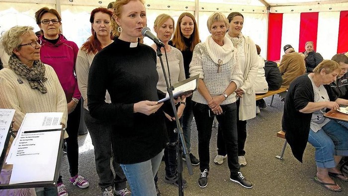 En præst holder et tale ved et indendørs arrangement, omgivet af en gruppe mennesker.