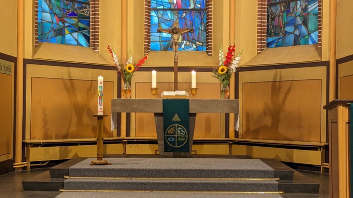 Jahreskerze, links stehend, vor Altar mit zwei Kerzen, zwei Sträußen in Vasen und mittig ein Kruzifix darauf. im Hintergrund drei Kirchenfensterausschnitte.