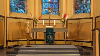 Jahreskerze, links stehend, vor Altar mit zwei Kerzen, zwei Sträußen in Vasen und mittig ein Kruzifix darauf. im Hintergrund drei Kirchenfensterausschnitte.