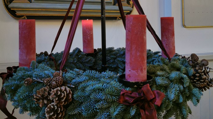 En adventskranse med røde lys og grønne grane foran en tavle med skrift