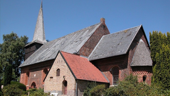 Kirchengebäude mit spitzem Turm und rotem Dach, umgeben von Grün.