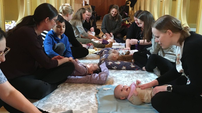 En gruppe af voksne og børn sidder på gulvtepper i en stor sal og spiser.