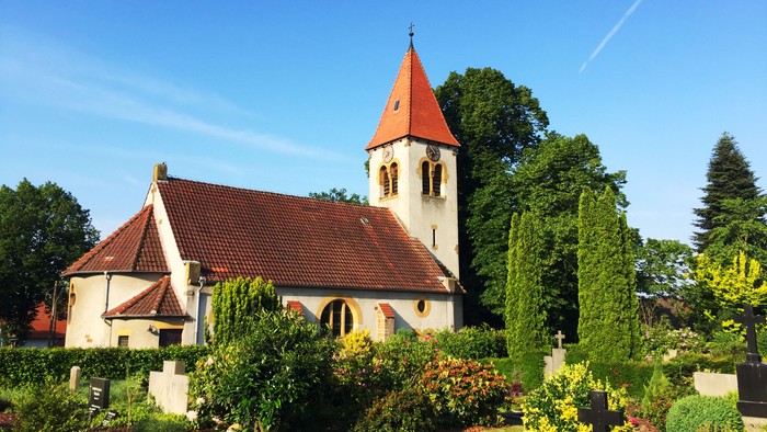 Kirchengarten mit weißem Kirchturm und roter Spitze, umgeben von üppigem Grün und blühenden Pflanzen