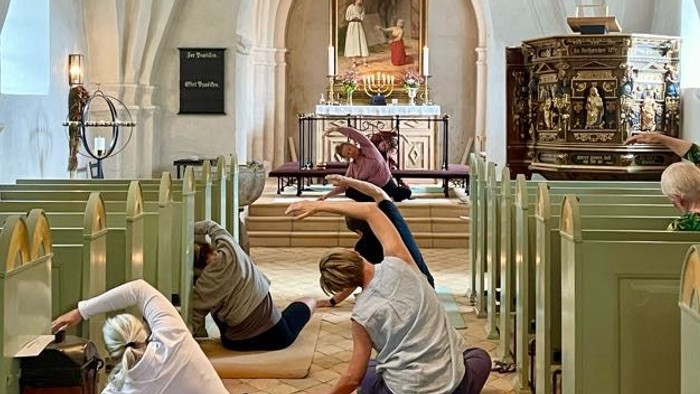 Mennesker laver yoga-strækøvelser inde i en historisk kirke med grønne kirkebænke og prægtige dekorationer.