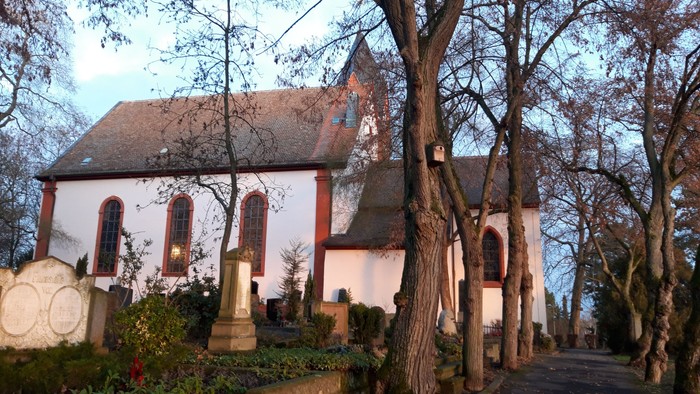 Kleine Kirche mit hohem Kirchturm, umgeben von Bäumen und Gräbern