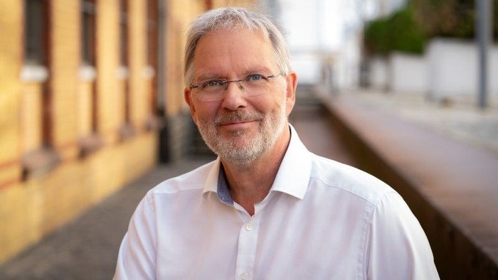 Ein lächelnder Mann mit Brille steht entspannt und freundlich im Freien in der Nähe eines Gebäudes.