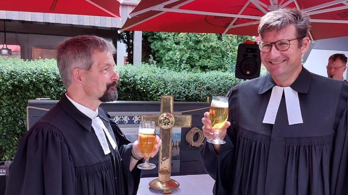 Zwei Männer in Richterroben stoßen mit Gläsern Bier bei einer Outdoor-Veranstaltung unter roten Schirmen an.