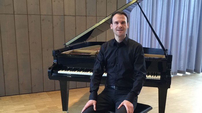 Ein Mann sitzt an einem Flügel in einem geräumigen Raum mit holzvertäfelten Wänden.