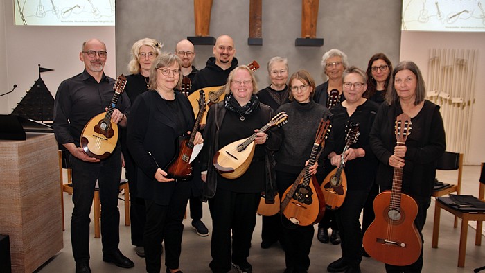 Eine Gruppe von Musiker:innen mit verschiedenen Saiteninstrumenten posiert gemeinsam drinnen für ein Wiedersehen oder eine Veranstaltung.