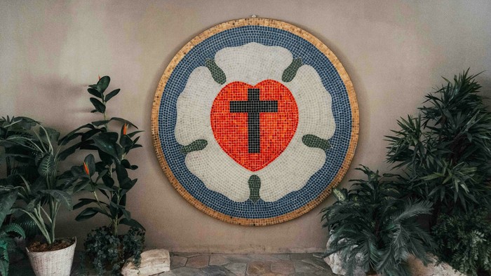 Ein Mosaikherz mit Kreuz an der Wand zwischen zwei Topfpflanzen