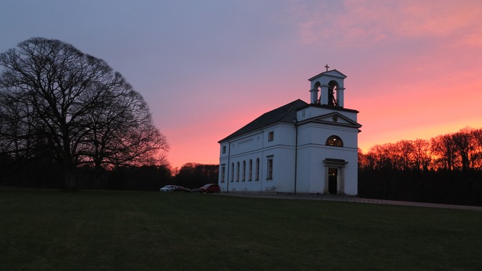 En hvid kirke med tårn og klokke ved solnedgang i en grøn og træfattig landskab