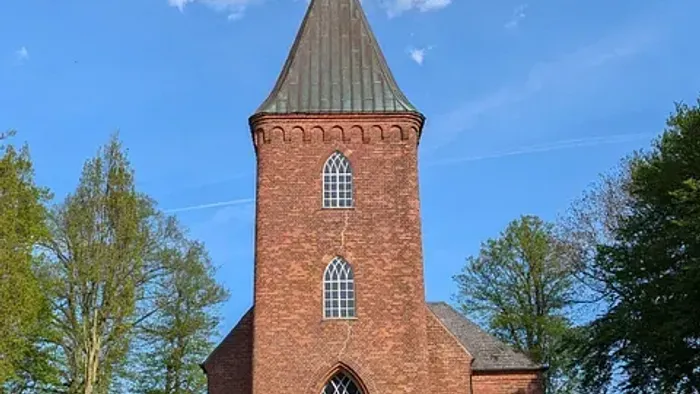 Kirchturm aus rotem Backstein vor blauem Himmel und Bäumen