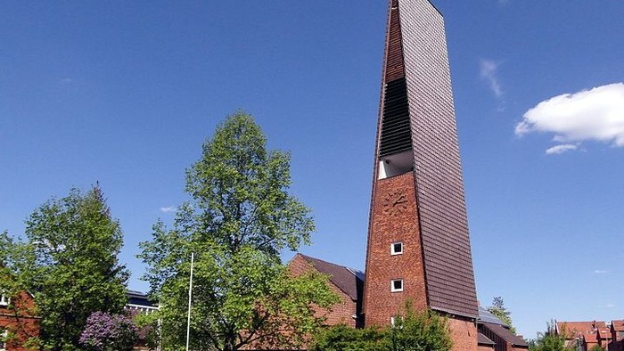Ein roter Backsteinturm mit spitzem Dach und Uhr, umgeben von Bäumen und Büschen, unter blauem Himmel