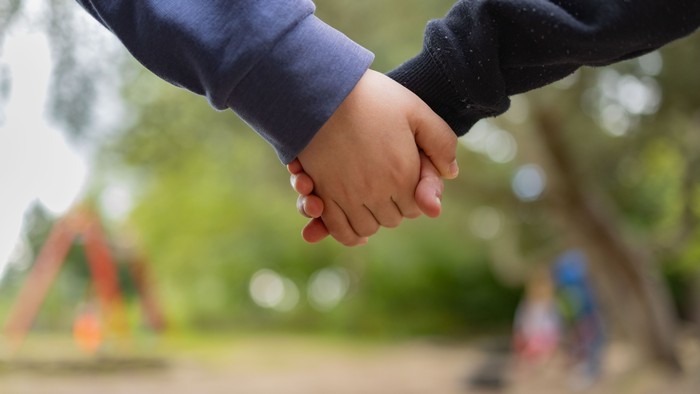 Zwei Menschen halten sich in einem Park an den Händen, umgeben von Grünflächen und Spielplatzgeräten.
