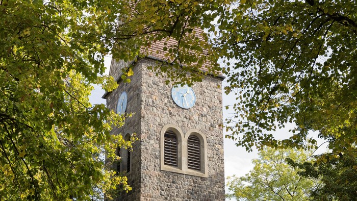 Der Kirchturm aus Stein mit Uhr und Fenster steht in einem Park.