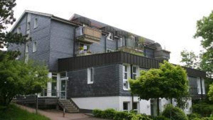 Das Bild zeigt ein zweistöckiges Haus mit grauer Fassade und weißen Fensterrahmen, umgeben von grünen Büschen und Bäumen.