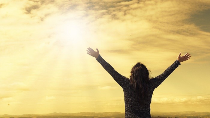 En person med udstrakt arme står på en højsæd og ser ud over landskabet mod solen.