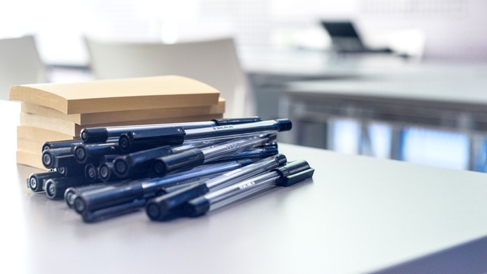 Flere sorte kuglepenne ligger spredt på et hvidt skrivebord nær en stak bøger.