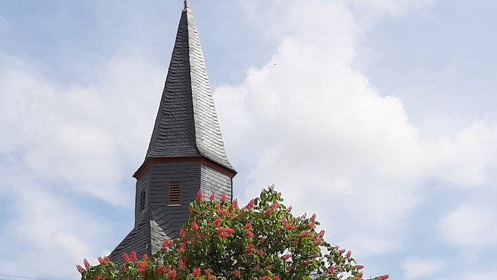 Ein blühender Baum vor einer malerischen Kirche mit hohem Kirchturm unter einem teilweise bewölkten Himmel.