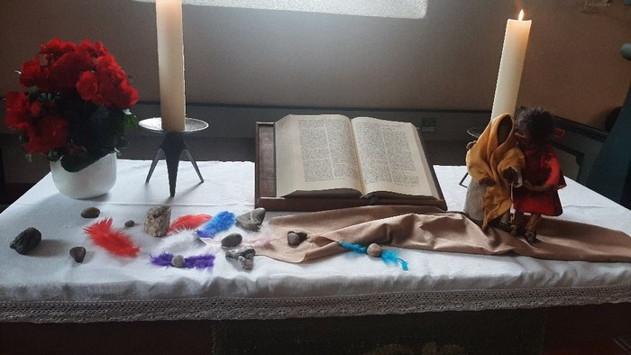 Eine ruhige Altargestaltung mit Kerzen, Blumen, einem offenen Buch und verstreuten bunten Federn auf einem bedeckten Tisch.