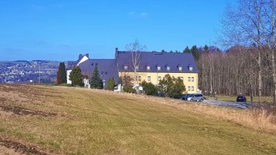 Ein großes gelbes Gebäude mit dunklem Dach auf einem Hügel, umgeben von Bäumen und Gras.