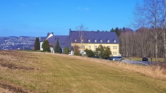 Ein großes gelbes Gebäude mit dunklem Dach auf einem Hügel, umgeben von Bäumen und Gras.
