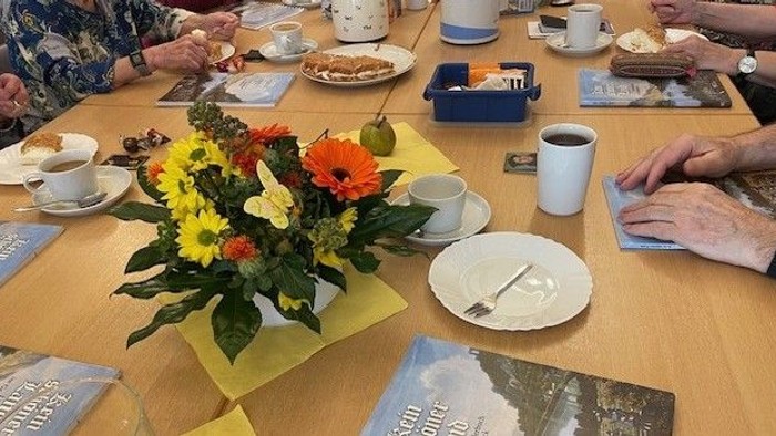 Eine Gruppe von Menschen trinkt Tee und isst Snacks, mit einer floralen Tischdekoration in der Mitte des Tisches.