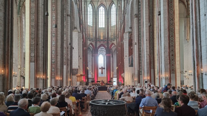 Große Menschenmenge in einer Kathedrale, viele sitzen auf Stühlen, hohe Decken, bunte Fenster.