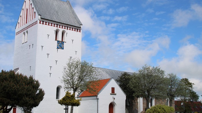 Hvid kirke med klokketårn og røde dørrer, omgivet af træer og blå himmel