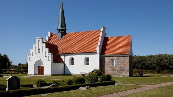 En hvid kirke med rødt tag og spir med tårn, omgivet af græs og træer