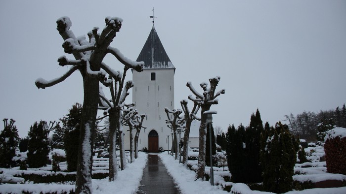 Der ses en våd flisegang, som fører hen til Nr. Løgum kirkes indgang i kirkens tårn. Der ligger sne ved siden af gangen, på de stynede lindetræer, på de små hække omkring gravstederne og lidt på kirketårnets tag. Himlen er grå